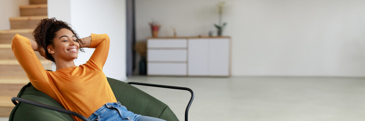 Rest Concept. Happy black lady sitting on comfortable bean bag at home in living room. Cheerful casual woman with closed eyes relaxing on sofa, leaning back, panorama with copy space