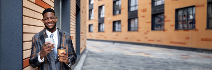 Wall Mural - Handsome black businessman in coat with coffee to go and smartphone standing next to office building wall, smiling to camera, having break after successful business meeting, copy space