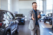 Salesperson at car dealership selling vehicles. Front view of handsome man standing in modern car center and posing. Bearded manager smiling and looking at camera.