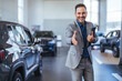 Good looking, cheerful and friendly salesman poses in a car salon or showroom and looks at camera. Successful businessman in a car dealership - sale of vehicles to customers
