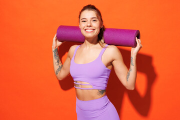 Wall Mural - Young confident happy fitness trainer instructor sporty woman sportsman wear purple top clothes spend time in home gym hold yoga mat isolated on plain orange background. Workout sport fit abs concept.