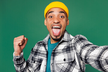 Wall Mural - Close up young man of African American ethnicity wear shirt blue t-shirt yellow hat doing selfie shot pov on mobile cell phone do winner gesture isolated on plain green background. Lifestyle concept.