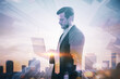Attractive young european businessman with laptop standing on blurry airy city background with mock up place. Future, tomorrow and success concept. Double exposure.