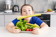 Happy little girl holding a stalk of celery in her mouth while holding a plate of fresh vegetables in front of her. Girl having fun in the kitchen at home. The concept of healthy nutrition.