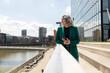 © Westend61 - Senior businesswoman holding mobile phone leaning on railing by river in city