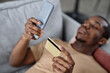 © AnnaStills - Young Black man relaxing on sofa at home paying for goods in online store with credit card, high angle shot