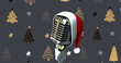Image of microphone with santa claus hat over trees on grey background
