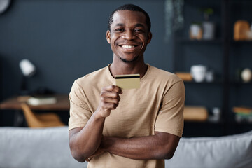 Medium portrait of cheerful young African American man standing in living room demonstrating bank card, copy space