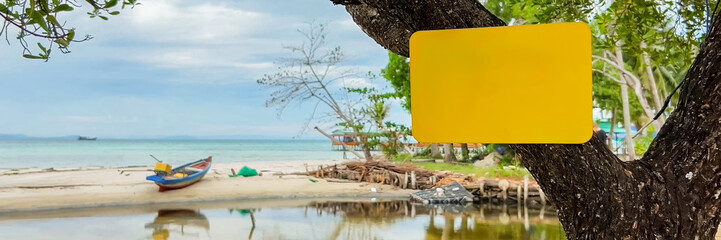 Wall Mural - Empty yellow sign hanging on a tropical tree with a serene beach, traditional boat, and clear sea in the background, ideal for summer vacation concepts