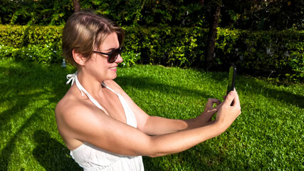 Wall Mural - Caucasian woman in sunglasses taking selfie with smartphone in tropical park, concept of summer vacations and World Tourism Day