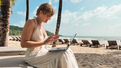 Wall Mural - Caucasian woman using a laptop on a tropical beach with palm trees, embodying leisure technology use and summer vacations, freelancer, digital nomad