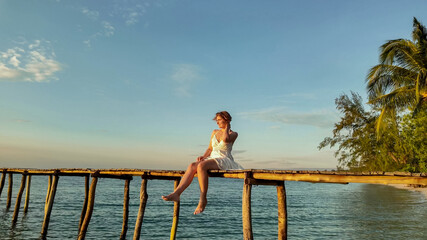 Wall Mural - Joyful Caucasian woman in white dress sitting on a tropical beach at sunset, expressing happiness and freedom, related to travel and summer vacations