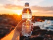 Hand holding a transparent plastic water bottle against a sunset backdrop with the sun shining through