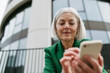 Mature businesswoman holding smartphone, waiting for business partner in the city. Beautiful older woman with gray hair standing on city street.