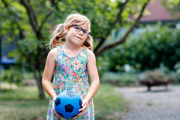 Wall Mural - Little preschool girl with eyeglasses playing with ball outdoors. Happy smiling child catching and throwing, laughing and making sports. Active leisure with children and kids. Summer day on backyard