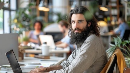 Poster - Stylish young man using laptop and monitor in marketing agency with colleagues in the background. Long-haired bearded manager seated at a desk in creative office. Fellow colleagues in the background.