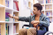 Side view portrait of young teenage boy with disability using wheelchair in library and choosing books on shelf