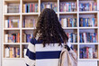 Back view of teenage girl with backpack choosing books on shelves in school library copy space