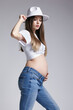 Young pretty pregnant woman in white t-shirt, hat and jeans. Female looking at pregnant belly. Belly exposed. 5th month of pregnancy