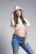 Young pretty pregnant woman in white t-shirt, hat and jeans. Female with belly exposed. 5th month of pregnancy