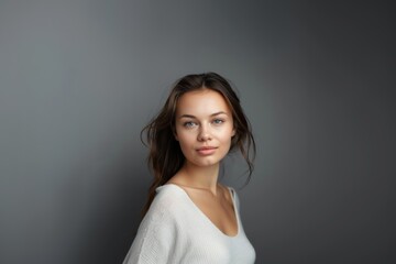 A brunette with wavy hair and a fleeting smile in light clothes on a gray background. Women's fashion and beauty, confident strong woman.