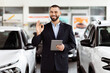 © Prostock-studio - A man standing in front of a car, holding a tablet in his hand. He appears to be inspecting something on the device, with the car parked behind him.