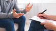 © Prostock-studio - A person is seated in a neutral-colored office space, engaging in a discussion with another individual who is taking notes on a pad, cropped