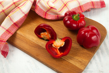 Cutting board with whole and half of red bell pepper and red kitchen towel on white wooden background. .