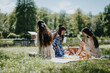 © qunica.com - Three sisters bonding over painting while enjoying a picnic on a sunny day in a scenic park, surrounded by greenery and a tranquil pond.