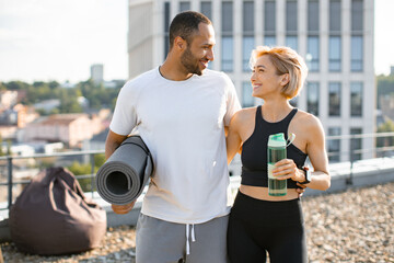 Wall Mural - Portrait of young couple doing sports outside house on urban background of city. Sporty woman standing with bottle of water and handsome man holding a mat.