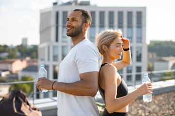 Wall Mural - Athletic fit female stands with her back to muscular sporty man and drinks water. Young couple engaged in physical education on terrace of high-rise building with urban city background.