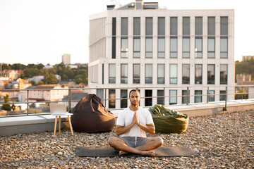 Wall Mural - Young athletic man doing yoga at sunset on urban background. Handsome male meditating in lotus position while sitting on mat on roof of modern house.