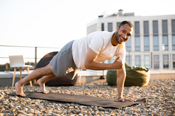 Wall Mural - Strong male in plank position on mat during sunrise against urban background. Young smiling sports man doing morning exercises on roof of modern house.