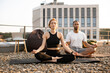 Young couple in sportswear sitting on mats in lotus position on roof of modern house. Athletic man and flexible woman meditating in morning on urban background.