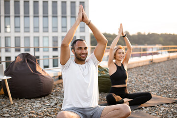 Wall Mural - Happy healthy couple meditating on roof on urban background in morning Physically strong man and woman sitting on mats and making yoga with hands raised above their heads. .