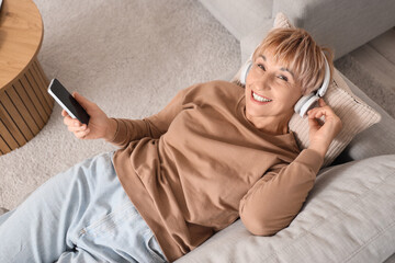 Poster - Mature woman in headphones with mobile phone listening to music on sofa at home, top view