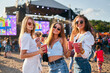 © artiemedvedev - Three women enjoy a sunny coastal music festival, sipping drinks, laughing, dancing to live beats. Casual beachwear, sunglasses, sunlit stage backdrop. Millennial summer party vibe by sea.