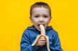 A small funny cheerful boy bites a banana, makes faces and plays around. Studio photography with yellow background.