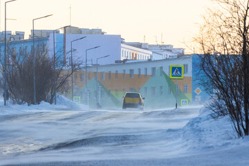 Wall Mural - A car is driving along a snowy street. View of the snow-covered roadway and sidewalk. Cold winter weather. Strong wind and blowing snow. The harsh northern climate of the polar region. Arctic.