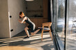 Side view of fit slim female doing single leg lunge using chair in living room. Attractive motivated fitness woman performing single leg lunge pose on chair. Home fitness and wellness concept