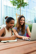 Two happy female college multiracial friends studying together in campus using laptop. Vertical image