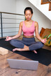 Young Asian woman meditating at home with online video meditation lesson using laptop. Vertical image.