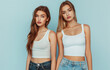 Two beautiful female models in their early twenties, one with long brown hair and the other with short blonde hair, wearing a white tank top and blue jeans