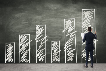 Poster - Back view of young european businessman drawing creative growing business graph sketch on black chalkboard wall. Financial growth, investment and success concept.
