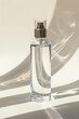 A transparent glass perfume bottle with a silver cap is placed on a white table. The bottle is half-full of a clear liquid. The background is a white wall with a soft shadow.