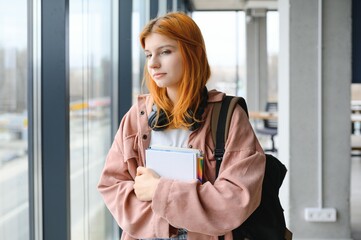 Wall Mural - Red-haired student girl stands by the window with books and a backpack