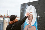 Fototapeta  - Female artist in black cap is painting picture with paint spray can spraying it onto canvas at outdoor street exhibition, side view of female art maker