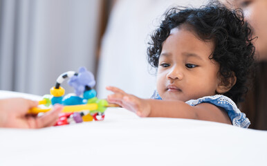 Portrait of adorable African 7 months old newborn baby girl with black curly hair standing near bed at home, looking and try to grab a colorful toy. Child care and development