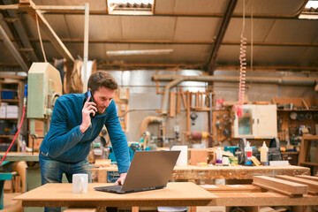 Carpenter Working In Woodwork Workshop Talking On Mobile Phone And Using Laptop