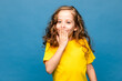 Happy little girl with curly hair is laughing covering her mouth with her hand on blue background.